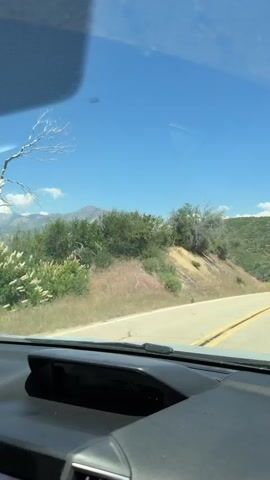 The video is captured from inside a car looking out through the windshield. The car is driving on a paved road that curves to the right. On the left side of the road, there is a dry, grassy hillside with some bushes and trees. Further in the distance, a large, rugged mountain range is visible under a clear blue sky with a few white clouds. The interior of the car, including the dashboard and a vent, is visible in the foreground. The time stamp on the video shows it is from 00:00 to 00:04.