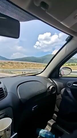 The video is filmed from the perspective of a passenger inside a car, looking forward. A car air freshener, orange and rectangular with a picture of a perfume bottle, hangs from the rearview mirror. In the foreground, the dashboard of the car is visible, including air vents and a car stereo. A disposable coffee cup is in a cup holder, and a plastic water bottle is next to it. Outside the car, a highway stretches into the distance, with rolling hills and fields on either side. The sky is blue with scattered clouds. The car appears to be moving on a sunny day.