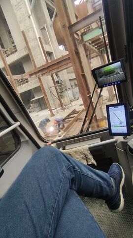 The video shows a view from inside a construction crane, likely from the operator's seat. Through the crane's window, a partially constructed building is visible, made of steel beams and concrete. Two construction workers are visible near the base of the structure, one of whom appears to be welding. Inside the crane cab, there are two small monitors mounted above the windshield. One monitor displays a live video feed of the crane's machinery. The other monitor shows a graph, potentially related to the crane's operational parameters. The lower portion of a person is also visible in the foreground, including their legs, clad in blue jeans, with a foot resting on the floor of the cab.