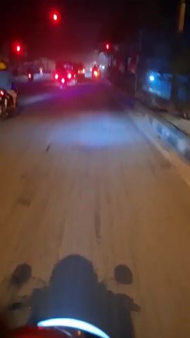 This is a first-person perspective video taken at night from a motorcycle. The rider is approaching a traffic light that is red. Several other vehicles, including motorcycles and cars, are stopped at the light. The road is paved and appears to be somewhat dusty. Trees and some flags are visible along the side of the road. The motorcycle's headlights are on, illuminating the road ahead and casting shadows of the rider and handlebars on the ground. The scene appears to be a city street or a busy intersection.