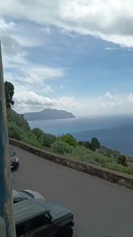 The video presents a panoramic coastal view in Genoa, Italy, captured from an elevated position. The scene opens looking out over the expansive, deep blue Ligurian Sea under a partly cloudy sky with patches of clear blue. A distant coastline or landmass is visible on the left horizon. The hillside below is covered with green trees and vegetation, dotted with houses that feature characteristic terracotta roofs. A dark-colored SUV is parked on the road in the immediate foreground.

As the camera slowly pans to the right, more of the intricate coastal landscape unfolds. The sky transitions from partly cloudy to mostly overcast with gray clouds. A large cargo ship can be observed moving across the distant sea. The hillside continues to be adorned with lush greenery and scattered residential buildings. A prominent feature along the road is a vibrant display of purple bougainvillea flowers cascading over a stone wall, adding a rich burst of color to the natural setting.

The pan continues to reveal more of the sloping terrain and the charming architecture of the houses nestled into the hillside. The sea remains visible, stretching out to the horizon. Towards the end of the video, a red SUV and a white car are seen parked on the curving road in the foreground. The overall impression is one of tranquil coastal beauty, showcasing the picturesque blend of sea, sky, and Ligurian architecture under a shifting sky. There is no visible text in the video.