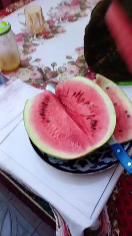 A hand holds a slice of watermelon, its pink flesh speckled with dark seeds, over a plate holding a halved watermelon. The halved watermelon is also pink with seeds and is sitting on a patterned plate. A knife with a blue handle is stuck into the halved watermelon. A glass jar with a yellow liquid and a green lid is on a tablecloth with a floral pattern, as is a glass.