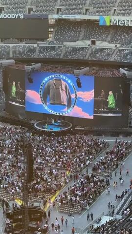 The video shows a large outdoor concert venue. A massive stage is set up with a large screen displaying abstract visuals and the performers. The screen shows a ring with a series of rotating images of clothing items and then two large red and pink balloons. Several performers in green outfits are on stage. In front of the stage, a large crowd of people are seated in tiered stadium seating, facing the stage. The stadium is filled with spectators. The overall scene depicts a live musical performance at a large stadium.