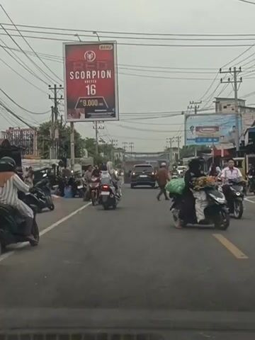 The video shows a busy street scene with numerous motorcycles and a few cars. Several people are riding on motorcycles, some with passengers. There are also pedestrians walking on the side of the road. Two large billboards are visible: one advertising "RED SCORPION 16" and another with text and images that are not clearly discernible. Power lines are strung across the sky. The overall environment appears to be urban or suburban, with buildings and shops lining the street. The sky is overcast. The video captures the movement of traffic and people, suggesting a normal day in a populated area.