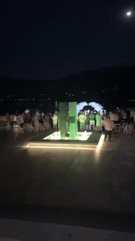A video shows a group of people gathered at night around a fountain. The fountain has changing colored lights, transitioning from green to blue. There are chairs set up in front of the fountain, suggesting an event like a ceremony or performance. Mountains are visible in the background, with lights from a distant town reflecting on the water.