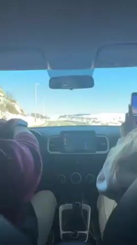 From the interior of a vehicle, the scene shows a driver and a passenger on a bright, sunny day, possibly winter, given the snowy landscape visible through the windshield. The driver, wearing a maroon long-sleeved shirt, has their hands on the steering wheel. The passenger is wearing a light-colored jacket and is holding a phone, seemingly taking pictures or videos. The vehicle's dashboard is visible, featuring a navigation screen, and there are several car accessories. The overall atmosphere appears casual and comfortable, as if the occupants are enjoying a scenic drive.