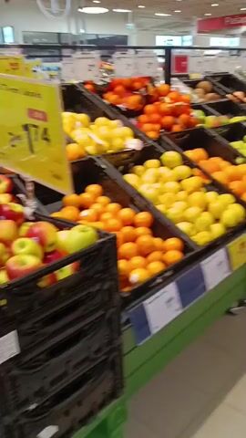 The video shows a wide shot of a grocery store produce section. In the foreground, there are several black bins filled with various fruits. From left to right, the bins contain green apples, oranges, lemons, pears, and kiwis. The fruits are neatly arranged in the bins, and some have price tags in front of them. In the background, there are more shelves and displays of produce, as well as signs and lighting typical of a supermarket. The overall impression is a well-stocked and organized fruit and vegetable display.