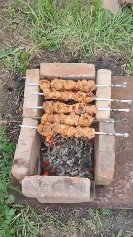 The video shows a shish kebab cooking over hot coals in a makeshift brick barbecue outdoors. Four metal skewers hold marinated pieces of meat, arranged horizontally above the coals. The fire is burning with visible flames amidst the charcoal. The barbecue is constructed of several red bricks laid out to form a rectangular pit. Green grass surrounds the barbecue area. The camera moves closer for a better view of the cooking meat.