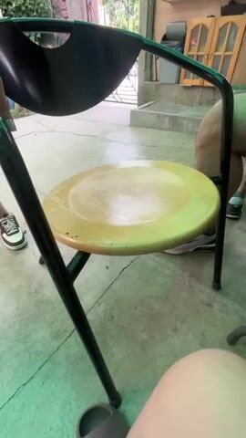 The video shows a person standing by a chair and a man sitting at a table. The person standing is wearing a white shirt and a black skirt, with long curly hair. They are holding onto the back of the chair. The man sitting is wearing a yellow shirt and has a beard. He is holding a cup and has his arm on the table, which is covered with a patterned tablecloth. There are several items on the table, including what appear to be packs of cigarettes and a small fan. The environment appears to be an outdoor or semi-outdoor area, possibly a patio, with a green corrugated roof overhead. There is also a glimpse of some cabinets in the background.