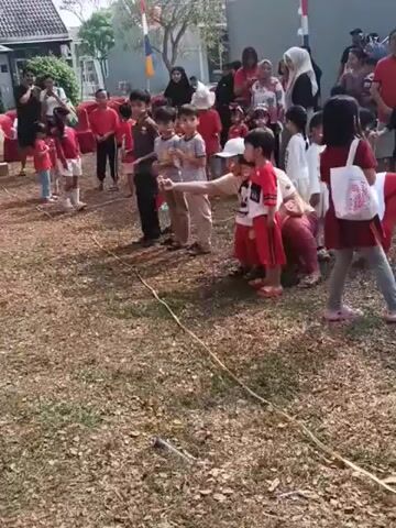 This video shows a group of children and adults gathered outdoors on a dry, grassy area. The children are mostly dressed in red and white clothing, suggesting a patriotic or festive occasion. Some children are lined up, possibly for a game or activity, with a yellow rope stretched out on the ground in front of them. An adult is kneeling down, interacting with some of the children. In the background, there are buildings and people observing the activity. The overall scene depicts a community event or a school activity.