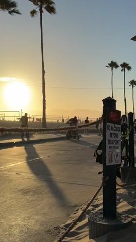 This is a vertical, eye-level, outdoor shot at dusk. The sun is setting on the left side of the frame, casting a warm, golden light across the scene. Palm trees line the background. In the foreground, a sandy beach is on the right, and a paved walkway runs through the middle. Several people are on the walkway, some on bicycles. A sign on the right reads "NO ALCOHOL BEYOND THIS POINT." A rope barrier separates the beach from the walkway. The overall atmosphere is relaxed and recreational.