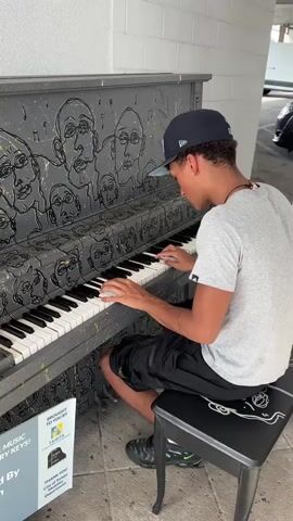 The video shows a young man playing a piano decorated with stylized face drawings in an indoor parking garage. He is wearing a light gray T-shirt, black shorts, black sneakers with green accents, and a dark blue baseball cap. He is seated on a black stool. The piano is painted gray with black line drawings of multiple faces on its surface. A sign next to the piano indicates it is part of a public art initiative. The parking garage has white walls and parked cars are visible in the background. The man is focused on playing the piano.