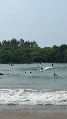 The video shows a group of people surfing in a tropical bay. In the foreground, waves break on a sandy beach.  A woman in a pink top and white hat is surfing towards the shore.  Several other people are either on surfboards or swimming in the water, some of them lying on their boards. In the background, a densely vegetated headland is visible. The sky is overcast.