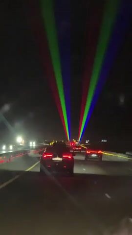 The video shows a nighttime drive on a highway. The perspective is from inside a vehicle, looking forward. Multiple cars are visible on the road ahead, with their taillights illuminated. Laser lights, in red, green, and blue, are projected upwards from a source ahead and spread out across the dark sky. The lasers appear to be part of a light show or event. The environment is dark, with the road marked by illuminated lines and barriers on the side. There is some fog or mist in the air, which catches the light. The cars are moving along the highway.