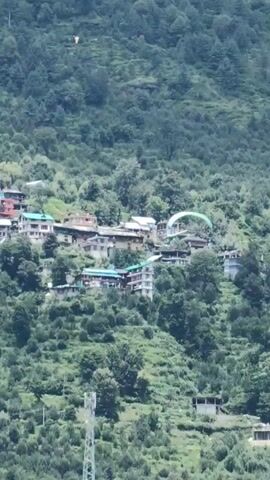 The video shows a village nestled on a lush green mountainside. The houses are clustered together, with some appearing to be multiple stories high.  There is a paraglider in the air, appearing to be in mid-flight over the village. The surrounding landscape is covered in dense foliage, with trees and vegetation dominating the slopes. In the foreground, there are rocks and a river or stream. A telecommunications tower is visible on the left side of the frame.