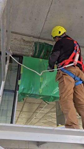 A construction worker wearing a yellow hard hat, safety glasses, dark jacket, brown cargo pants, and a safety harness stands on a silver platform. He is facing a concrete ceiling where green pipes are visible. A green plastic sheet is taped to the ceiling near the pipes. The worker uses a small hammer and chisel to work on the concrete ceiling. The environment suggests an unfinished or under-construction building. There's a large window behind the worker, offering a view of other construction or building elements. The lighting is natural and bright.