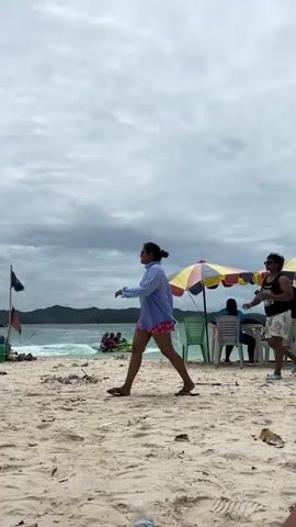 Two individuals walk across a sandy beach. The person on the left is wearing a blue button-down shirt and red shorts and is holding a phone. The person on the right is wearing a black tank top and patterned shorts and is carrying a backpack. In the background, there is the ocean with choppy waves, a jet ski, several boats docked, a few beach umbrellas, and distant islands under a cloudy sky.
