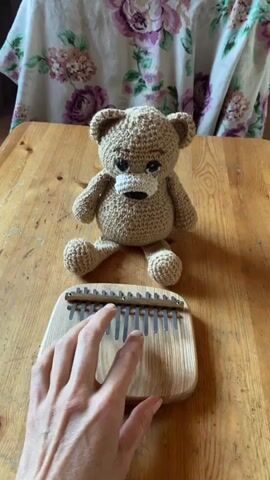 A hand plays a kalimba, a wooden instrument with metal tines, which is positioned on a wooden table. A crocheted teddy bear sits behind the kalimba on the table, facing the camera. The background features a floral fabric draped behind the bear. The lighting appears to be natural.