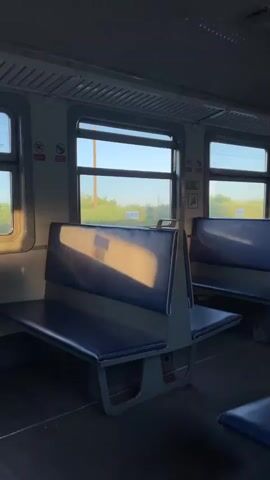This video shows the interior of a train carriage, likely in Russia, given the location context of Kiselyovsk. The primary subjects are the empty blue vinyl seats and the windows of the train. The scene is set within a typical passenger train car, with the seats arranged facing each other, separated by a central aisle.

The activity taking place is the train in motion, as evidenced by the passing scenery visible through the windows. The sunlight streaming in suggests it is either early morning or late afternoon, casting long shadows across the seats and floor. The weather appears to be fair, with a clear sky and greenery visible outside. There are no people visible in the carriage.

Notable details include the utilitarian design of the seats, the ribbed ceiling panels, and the large windows offering views of fields and trees. On the wall above the windows, there are some signs. One sign, to the left, appears to have a cigarette with a diagonal line through it, indicating "no smoking." Another sign, to the right, shows a person in a wheelchair, suggesting accessibility. The overall mood is peaceful and contemplative, perhaps evoking a sense of journey or solitude. The specific location cues are limited to the general appearance of the train interior and the countryside outside, which is consistent with a rural or semi-rural setting in Russia.