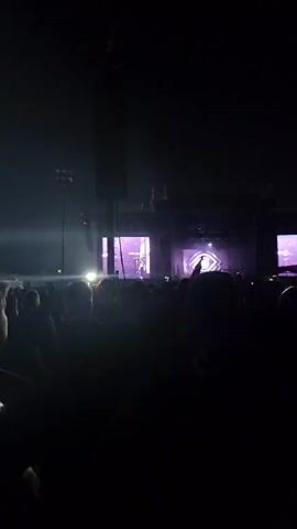 The video shows a concert scene, filmed from within the audience. The view is mostly obscured by the backs of people's heads, but in the background, a stage is visible, illuminated with purple and white lights. There's a large video screen on the left side of the stage showing a live feed of the band.  A person is holding up a cell phone, recording the performance. The video quality is a bit shaky and the lighting is dim due to the concert setting. The band is playing, and the music has a heavy rock sound.
