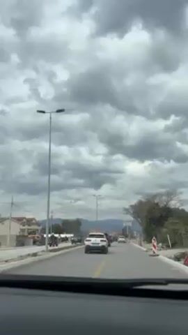 The video shows a view from inside a car driving on a road. There are cars in front, including a white Citroën. Streetlights line the road, and construction appears to be underway on the side. The sky is overcast with dark, heavy clouds. Distant mountains are visible in the background. A sound like a car horn can be heard.
