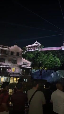 At night, a group of people is watching a light show in front of a traditional Chinese pagoda and wall. The pagoda, illuminated in pink, stands on top of a hill covered in green trees. Beams of light emanate from the pagoda, reaching towards the sky. Below the pagoda, the stone wall is also illuminated with white light, creating a visual display. The crowd of people stands in the foreground, their backs facing the camera as they observe the scene. There are buildings visible on the left side of the frame. The sky is dark, and the lights add vibrancy to the environment.