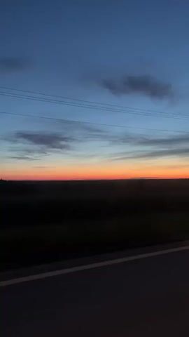 The video shows a view from a car driving alongside a road at dusk or dawn. The sky is a gradient of blue to orange, with some clouds visible. There are power lines running across the sky. Below the horizon is a dark, indistinct area, likely fields or trees. The car's dashboard or window frame is visible in the foreground. Music plays throughout the video.
