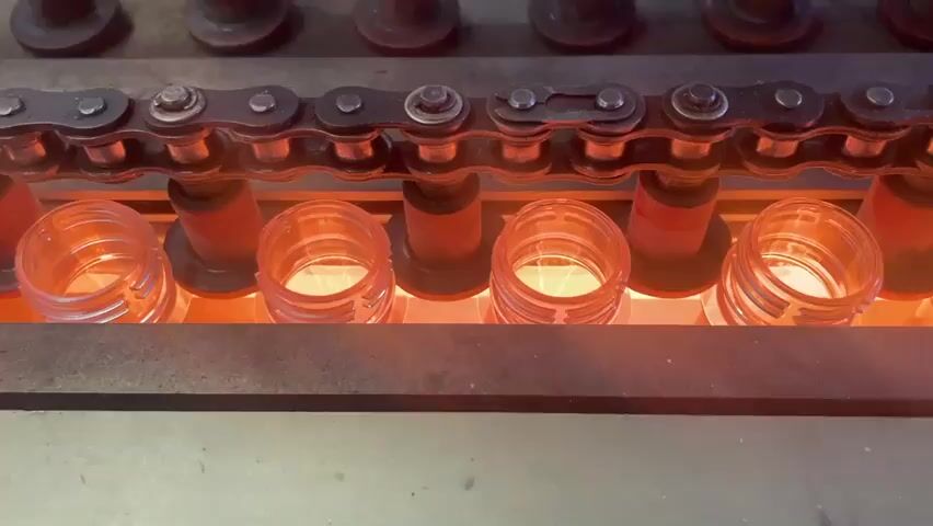 The video shows a close-up view of a conveyor belt system in a manufacturing facility. A chain conveyor moves several clear plastic bottle preforms through an area illuminated by red heat lamps. The preforms appear to be in an inverted position, with their open ends facing upwards, being heated from below. The heat lamps cast a warm orange glow on the preforms and the surrounding machinery, which includes metal components and wooden or particle board fixtures. The preforms are arranged in a single line, spaced evenly along the conveyor. The process seems to be part of a bottle manufacturing or filling line, where the plastic is being softened or prepared for further processing. The environment appears to be industrial, with a focus on automated machinery.