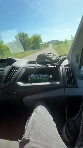 From the perspective of someone in the driver's seat, the video shows a car driving on a country road with green trees and grass on either side. The sky is blue and there's a building in the distance. Inside the car, the dashboard is visible, with a bottle of green liquid and a bag in the storage area. The person driving is wearing jeans. Music is playing.