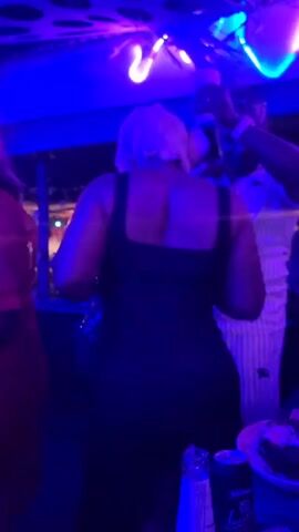 The video shows a woman in a black dress dancing in what appears to be a nightclub or bar, characterized by blue lighting and a neon flamingo sign overhead. A man in a striped shirt stands to her right, holding a drink. There's a table in the foreground with bottles and a plate of food. Other figures can be seen in the background. The woman's back is to the camera, and she has short, light-colored hair. The overall atmosphere is lively and suggestive of a social gathering.