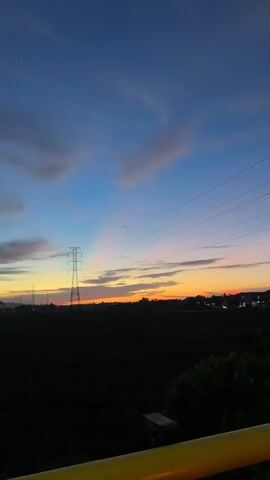 This media file captures a scenic view of a sunset in Meureudu, Indonesia. The primary subjects are the striking silhouette of a tall electrical transmission tower and the vibrant hues of the twilight sky. The scene is set in a rural or semi-rural landscape, characterized by dark, silhouetted fields or vegetation in the foreground and middle ground. In the distance, a faint line of buildings and more transmission towers suggest a developing area or the outskirts of a town. The sky is a masterpiece of nature, transitioning from a deep blue at the top to soft oranges and yellows near the horizon, where the sun has just set. Wispy clouds are scattered across the sky, catching the fading sunlight and adding texture and depth to the scene. The overall mood is one of tranquility and natural beauty, enhanced by the dramatic contrast between the dark landscape and the illuminated sky. The time of day is clearly dusk, as indicated by the sunset colors and the lengthening shadows. The presence of electrical infrastructure suggests a populated area, but the focus remains on the natural spectacle. No visible text is present in the media file.