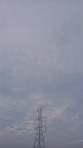 A low-angle shot of a transmission tower and power lines against a cloudy sky. The tower is metallic and lattice-style, with multiple levels of crossbars. The sky is overcast with a uniform grey hue, suggesting a dull or possibly rainy day. The image is static, with no discernible movement.