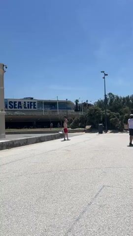 The video shows an outdoor scene on a paved surface under a clear blue sky. There are two young men visible. The first man, wearing red shorts, is on a skateboard in the left half of the frame, moving from left to right. The second man, wearing black shorts and a white tank top, is further to the right, walking away from the camera. In the background, there is a building with "SEA LIFE" written on it, a basketball hoop, trees, and streetlights. The scene appears to be a public area, possibly a park or recreational space near the coast.