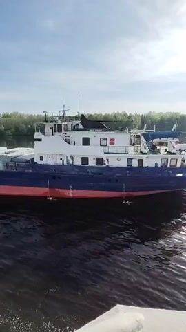 The video shows a river with a long, two-story boat moving on the water. The boat has a white top portion, a dark blue middle, and a red stripe near the water. Green trees line the river bank in the background under a sky with scattered clouds. The camera appears to be on another boat moving alongside it, visible by a white portion on the left of the frame. The word "Пайла" is written in white letters on the boat's side.