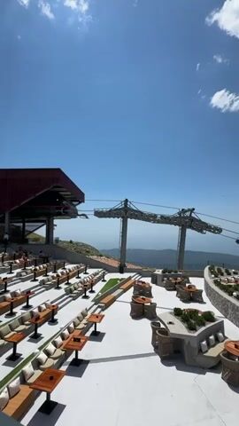 This video pans across a rooftop dining area with a clear blue sky and scattered clouds above. In the foreground, there are numerous outdoor tables and seating arrangements, including lounge-style benches with cushions and individual tables. Some tables are set up in rows, while others are grouped around stone planters filled with greenery. In the middle ground, there are more seating areas and an outdoor bar or serving station. In the background, a cable car system is visible, with large metal towers supporting the cables that extend across the landscape. The area appears to be situated at a high elevation, with a vast, hazy landscape visible in the distance. There are a few people visible in the far left of the frame, near the cable car station. The overall scene suggests a resort or restaurant with a panoramic view.