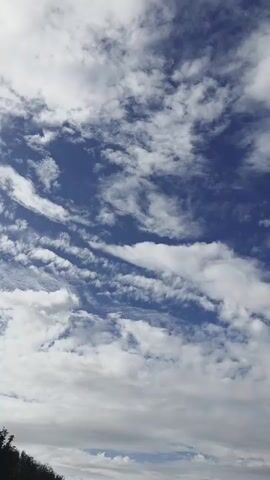 The video shows a sky filled with clouds. The clouds are white and fluffy, with some appearing wispy. The sky is blue, indicating it is daytime. The bottom left of the frame shows the tops of dark green trees.