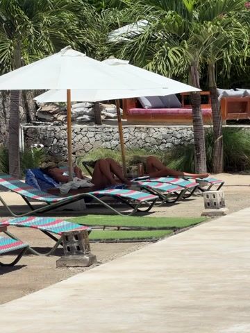 This outdoor, eye-level shot captures a serene resort or beach club setting in Kecamatan Nusa Penida, Indonesia, on a bright, sunny day. The scene features two individuals, likely women, relaxing on colorful lounge chairs beneath two large white umbrellas, seeking respite from the sun. The person on the left is partially visible, wearing what appears to be swimwear and looking down at a phone or small device. The second person, further right, is also lying back on a lounge chair, with their body mostly obscured by shadow and the chair itself.

The foreground shows more empty lounge chairs, suggesting a spacious area. Underneath some of the chairs, patches of artificial green turf are laid out on the sandy ground, next to decorative stone lanterns or stands. A concrete pathway runs along the right side of the frame. In the background, lush green palm trees frame the area, indicating a tropical environment. Behind the umbrellas, a stone wall is visible, with a cushioned outdoor seating area further back, adding to the luxurious resort ambiance. The bright light and clear shadows suggest it is mid-day or early afternoon, contributing to a relaxed and leisurely atmosphere. No discernible text is visible in the image.