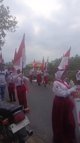 A group of children in red and white uniforms, holding red and white flags, are marching in a procession along a paved road. An adult male in a blue and white shirt and jeans walks alongside the children. The sky is overcast. There is a motorcycle parked on the left side of the road. The flags appear to be national flags. The children are wearing hats and sashes. Some of the children are carrying poles with the flags. The scene suggests a parade or a patriotic event.
