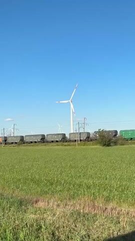 A train carrying cargo cars moves across a green field under a clear blue sky. In the background, several wind turbines are visible, with one prominent turbine in the center of the frame. The camera perspective is from inside a vehicle, with a rearview mirror visible in the lower corner of the frame.