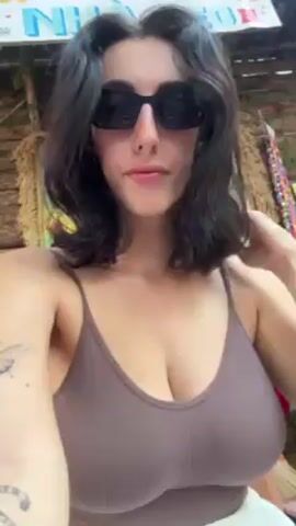 A woman wearing a brown tank top, a white skirt, and black sunglasses is sitting on a wooden chair outdoors. She is facing the camera and runs her fingers through her dark, shoulder-length hair. The camera pans down to show her lower body, and reveals that she is surrounded by colorful round decorations resembling pom-poms on the ground. Some decorations are red, yellow, green, and pink. Her feet are visible and she is wearing light-colored shoes. The background includes a painting, some colorful decor, and what appears to be a textured wall. The overall setting appears to be casual and sunny.