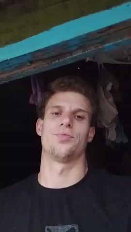 The video is a close-up selfie of a young man with curly brown hair and a short beard, wearing a black t-shirt. He is looking directly at the camera and smirking. The background is dark, with some blurred objects hanging from what appears to be a wooden structure overhead. The lighting is dim, suggesting an indoor or nighttime setting. The man's expression is playful and slightly mischievous. There are no other people or significant objects in the frame. The video appears to be a short, casual clip.