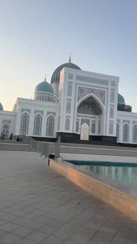 The video opens with a wide shot of a large, ornate white mosque with turquoise domes. The sun is low in the sky, casting a warm glow on the building and the surrounding plaza. In the foreground, there is a tiled area and a large, rectangular reflecting pool. The camera pans to the right, revealing more of the mosque's facade, which is decorated with intricate geometric patterns. As the camera continues to move, a tall, slender minaret comes into view. The sky is a pale blue, with a few wispy clouds. The overall impression is one of peace and tranquility. There are no people visible in the immediate vicinity of the mosque, though there appear to be a few figures in the distance near the entrance. The environment seems to be an open, public space, possibly a park or plaza, designed to complement the grandeur of the mosque. The water in the reflecting pool shows a clear reflection of the sky and the surrounding structures.