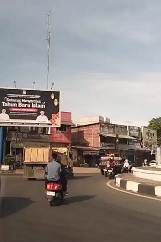 A street scene is shown with various vehicles and buildings. A billboard with an Islamic greeting for the new year is prominent. Several motorcycles and a truck are visible on the road. People are riding the motorcycles, one wearing a headscarf. Buildings with multiple stories line the street, appearing to be shops or businesses. The sky is clear with a few clouds. The scene suggests a busy urban or semi-urban environment.
