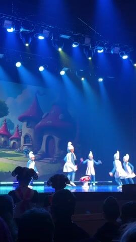 Five young girls perform a dance routine on a stage, dressed as Smurfs in white dresses, blue tights, and white Smurf hats. The stage is illuminated by blue lights, and a cartoon backdrop of Smurf houses is displayed behind them. In the foreground, the heads and shoulders of audience members are visible, silhouetted against the bright stage.