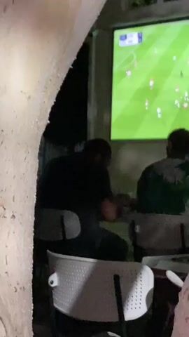 The video shows two men sitting at a table, facing away from the camera, watching a soccer game projected onto a large screen. The game is in progress, with players in white and red jerseys visible on the green field. The men appear to be in an outdoor setting, possibly a patio or restaurant, with the night sky visible through a window in the background. One man is wearing a black t-shirt, and the other is wearing a green and white patterned shirt. They are both seated on chairs and appear to be engaged in watching the game. The projection screen is the focal point of their attention.