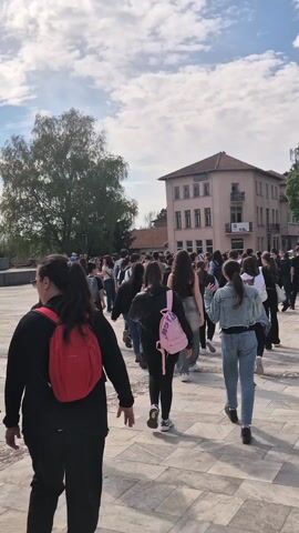 The video shows a large group of people walking across a large, paved plaza. The people appear to be mostly young adults or teenagers. They are wearing various types of casual clothing. Many are carrying backpacks. The plaza is surrounded by buildings, including a three-story, pinkish-beige building with a brown roof. There are also several large trees. The sky is partly cloudy. The video is taken from the perspective of someone walking with the crowd. The sound of people talking can be heard in the background.