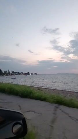 The video shows a view from a vehicle driving along a paved road next to a body of water. On the left side of the frame, a side-view mirror is visible, reflecting a dark interior. The road surface appears worn with some patches. Beyond the road, there is a strip of green grass and then a sandy beach bordering the water. The water is choppy with small waves and reflects the sky. Several small boats are visible on the water near the shore, and in the distance, a thin line of land with some buildings can be seen. The sky is overcast with soft clouds, and the light suggests either dawn or dusk, with a hint of pink and orange in the sky. A large tree with green foliage is on the left side of the road in the latter half of the video, with a park bench visible beneath it. The overall impression is a peaceful coastal drive.