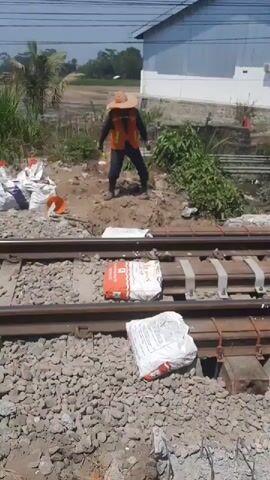 This video shows two construction workers near railway tracks. In the foreground, there are sacks of what appears to be cement next to the tracks. The workers are wearing safety vests and hats. One worker is hammering on the railway tracks, while the other watches him. In the background, there are buildings and vegetation. The scene suggests ongoing construction or maintenance work on the railway line.