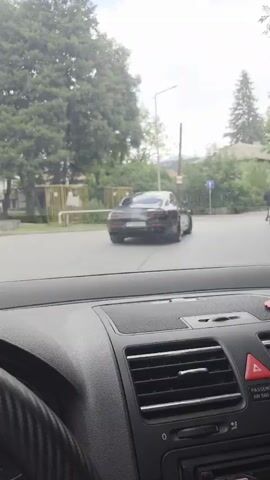 This is a first-person video taken from the interior of a car, looking out through the windshield. The car is in motion, suggesting it's a dashcam recording or a video taken by a passenger.

The scene is an urban or semi-urban environment, possibly a town or village, with buildings and trees visible. The road appears to be paved, with a crosswalk and traffic signs. The weather seems overcast, with a cloudy sky. The time of day is likely daytime, given the lighting conditions.

The primary subjects in the video are:

*   **A dark-colored car**: This car is in front of the camera's vehicle and is turning a corner. It appears to be a sedan or coupe.
*   **A cyclist**: A person on a bicycle is visible on the right side of the road, also in motion, riding in the same direction as the car.
*   **The interior of the camera's car**: Parts of the steering wheel, dashboard, and air vents are visible in the foreground. A speedometer is partially visible on the left, indicating the car's speed, but no specific numerical value can be read clearly.

The activity taking place is a vehicle traveling on a road, navigating through an intersection. There's a sense of everyday movement and traffic.

Notable details include:

*   **Location cues**: While not explicitly stated in the video itself, external context identifies the location as Razlog, Bulgaria. This suggests the architecture and natural environment might be typical of that region.
*   **Road infrastructure**: A blue and white road sign with a symbol is visible, likely a traffic regulation sign. A metal railing and a textured wall are also present on the left side of the road.
*   **Vegetation**: Lush green trees and foliage suggest it's likely spring or summer.

There is no discernible text within the visual frame of the video that provides specific information like place names or advertisements. The overall mood conveyed by the overcast sky and steady movement is neutral and observational.