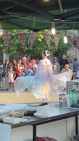 A young woman wearing a flowing white dress and a bun hairstyle with a white flower is dancing on a stage. Her dress has iridescent panels that catch the light. She is holding large, white, pleated wings that extend from her arms and back. She is performing in front of an audience of adults and children. Behind the audience, there are trees and a fence. The stage is covered by a green canopy, and colorful balloon flags are strung across the top. In the foreground, there is a table with various items, including containers, dishes, and bottles of liquid. The scene appears to be an outdoor event or festival.